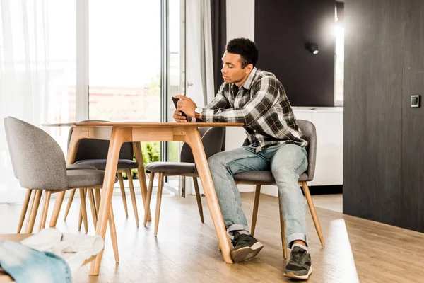 Full length view of african american man sitting before table and using smartphone — Stock Photo