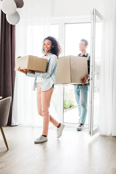 Full length view of african american husband and wife smiling while walking into house, holding boxes and looking at camera — Stock Photo