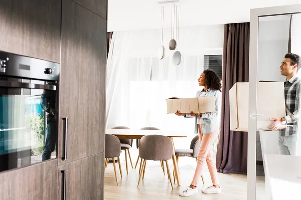 Vista de longitud completa del marido y la esposa afroamericanos sosteniendo cajas y mirando a la habitación mientras camina en casa - foto de stock