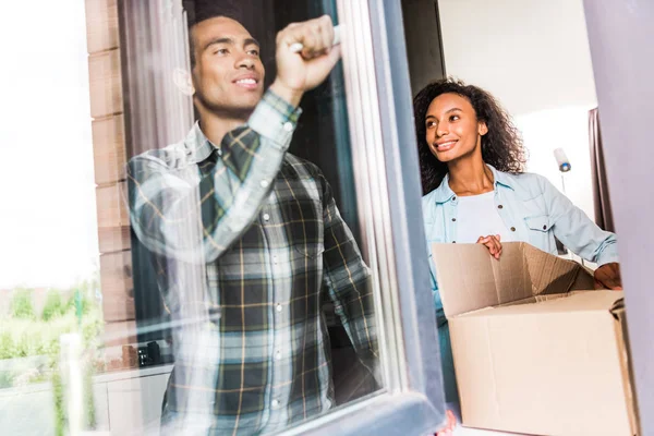 African american husband opening window while wife opening box and looking at husband — Stock Photo