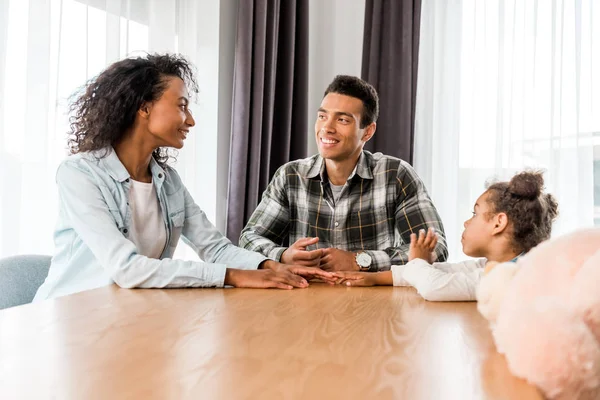 Familia afroamericana sentada frente a la mesa y sonriendo mientras los padres se miran - foto de stock