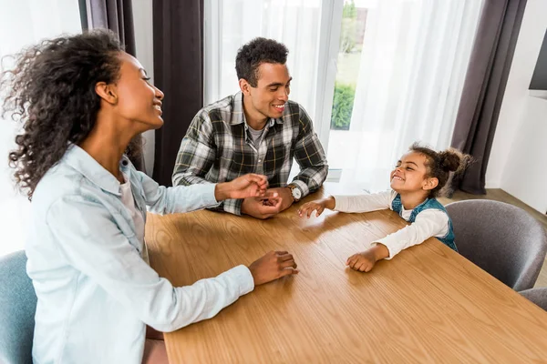Familia afroamericana sentada frente a la mesa, riéndose y mirándose - foto de stock