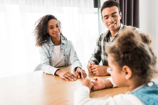 Selektiver Fokus der afrikanisch-amerikanischen Familie, die vor dem Tisch sitzt, während die Frau in die Kamera schaut und der Vater auf das Kind schaut — Stockfoto