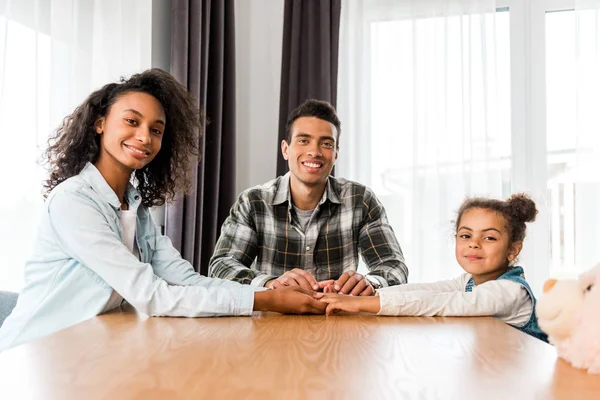 Familia afroamericana sentada frente a la mesa, sonriendo y mirando a la cámara - foto de stock
