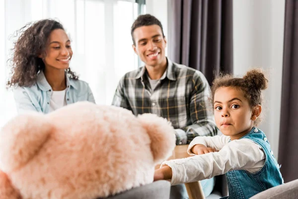 Selektiver Fokus der afrikanisch-amerikanischen Familie, die vor dem Tisch sitzt, während das Kind in die Kamera schaut und der Elternteil das Kind ansieht — Stockfoto