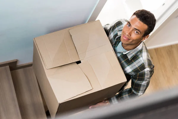 Vista de ángulo alto del hombre afroamericano guapo que sostiene la caja mientras que va arriba y mira la cámara - foto de stock