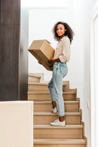 Full length view of african american woman walking upstairs while holding box and looking at camera — Stock Photo