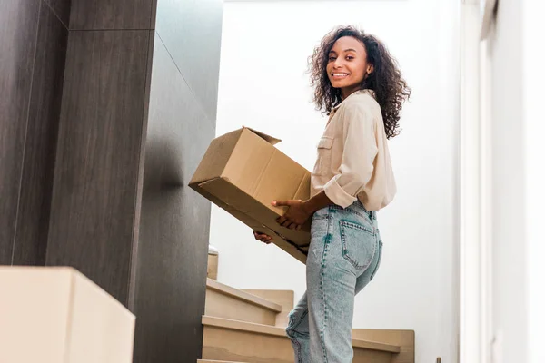 African american woman walking upstairs while holding box and looking at camera — Stock Photo
