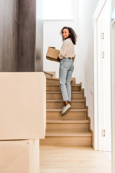 Full length view of beautiful african american woman walking upstairs while holding box and looking at camera — Stock Photo