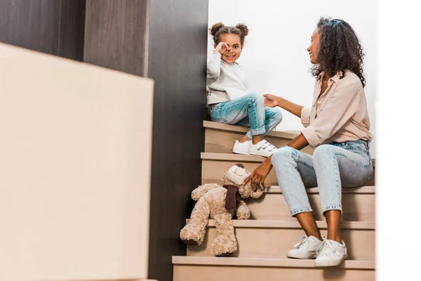 Full length view of african american woman with kid sitting on stairs while daughter smiling and looking at camera — Stock Photo