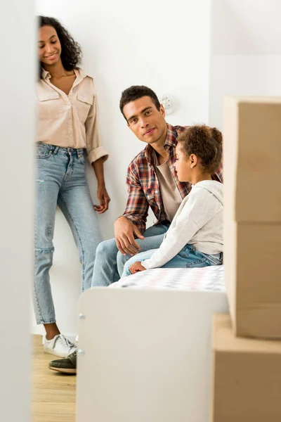Full length view of african american woman standing near wall while man sitting with kid on bed — Stock Photo