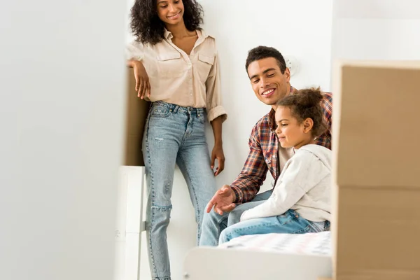 Messa a fuoco selettiva della donna afro-americana in piedi vicino al muro mentre l'uomo seduto con il bambino sul letto — Foto stock