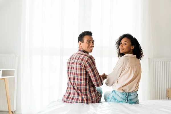 Africano americano guapo marido y esposa sentado en cama, cogido de la mano y mirando cámara - foto de stock