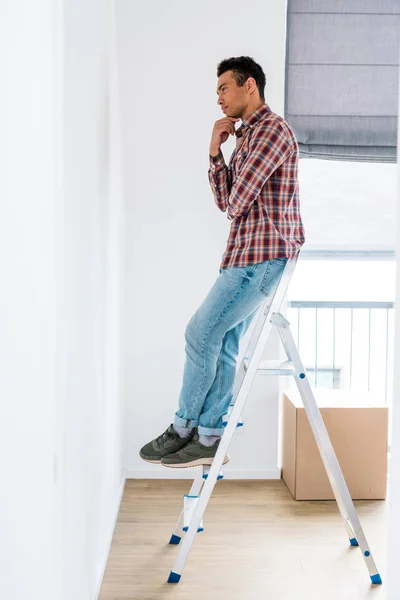Full length view of handsome african american man standing at ladder and looking at wall — Stock Photo