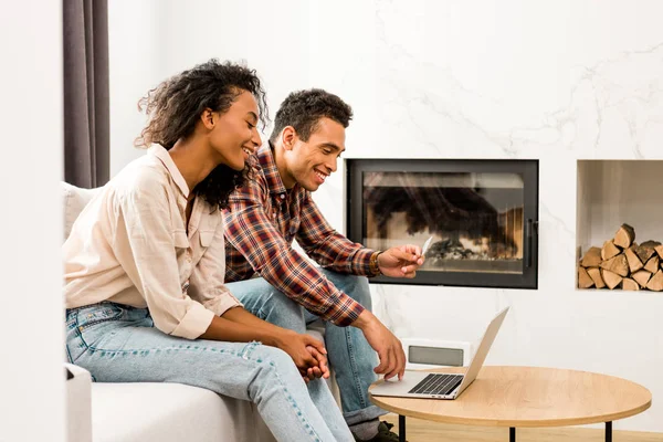 Vista lateral de la pareja sentada en el sofá y mirando a la computadora portátil mientras el hombre sostiene la tarjeta de crédito - foto de stock