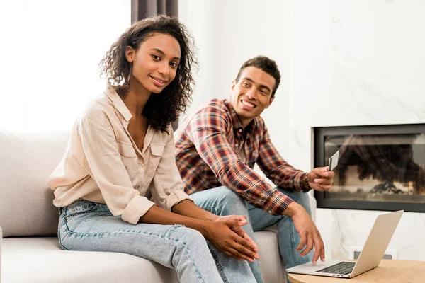 Africano americano pareja sentado en sofá mientras hombre celebración de crédito tarjeta y mujer mirando cámara - foto de stock