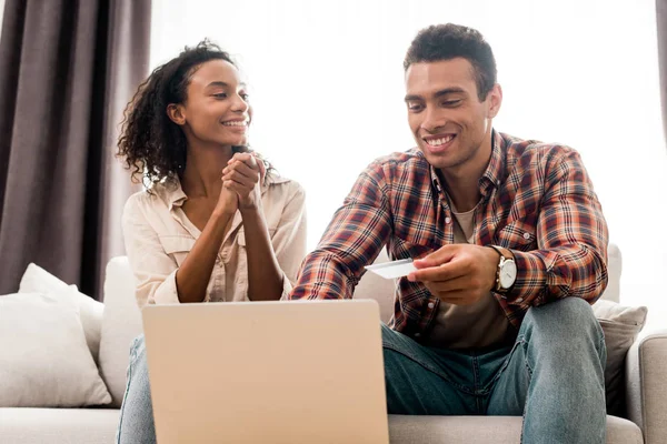 Handsome man and african american woman sitting on sofa while husband looking at laptop and holding credit card and wife looking at man with clenched hands — Stock Photo