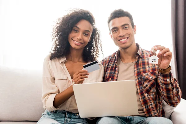 Front view of handsome african american man and woman looking at camera and sitting on sofa while wife holding credit card and husband holding key — Stock Photo