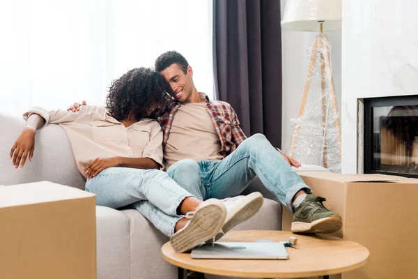 Vue pleine longueur de la femme afro-américaine et mari relaxant tout en étreignant et assis sur le canapé — Photo de stock