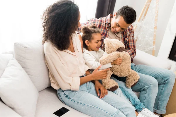 Africano americano familia sentado en sofá y sonriendo mientras padre jugando con hija - foto de stock