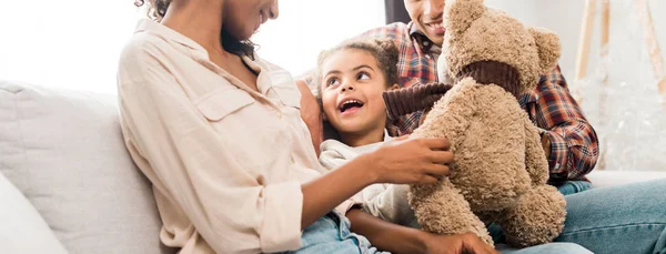 Panoramic shot of african american family sitting on sofa while kid holding teddy bear and looking at mother — Stock Photo