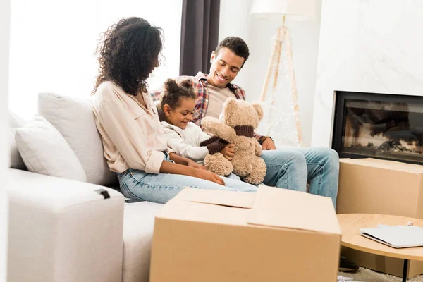 Афроамериканець сім'я сидить на дивані і посміхається в той час як дочка грає з плюшевого ведмедика і батьки дивляться на дитину — стокове фото