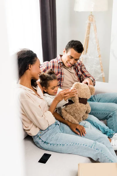 Enfoque selectivo de la familia afroamericana sentada en el sofá y sonriendo mientras su hija juega con el oso de peluche y los padres mirando al niño - foto de stock