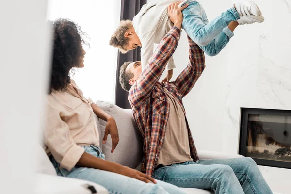 Selective focus of african american mother looking at dad while man holding kid in air — Stock Photo