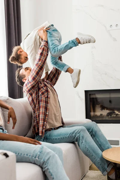 Cropped view of mother sitting on sofa near african american dad holding kid in air — Stock Photo