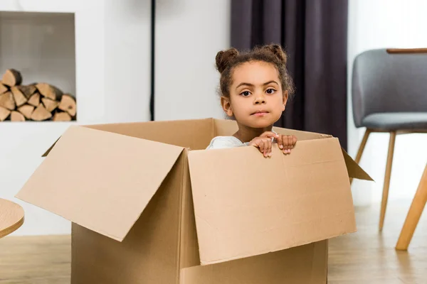 Cute african american child sitting in box and looking at camera — Stock Photo