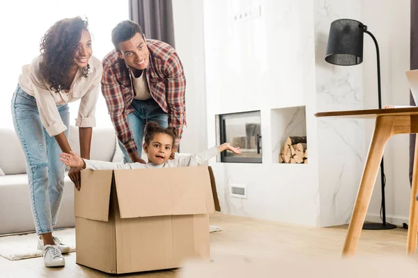 Vista completa de los padres afroamericanos jugando con la hija moviendo la caja con el niño - foto de stock