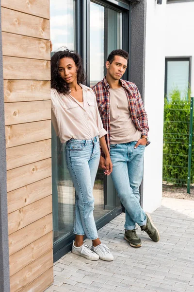 Visão de comprimento total do homem e da mulher americanos africanos inclinados na porta enquanto estão ao ar livre e olhando para a câmera — Fotografia de Stock