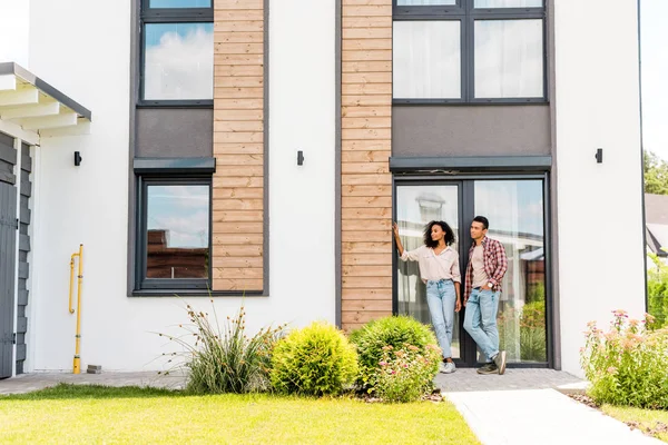 Full length view of african american wife and husband leaning on door while standing outdoors and looking away — Stock Photo