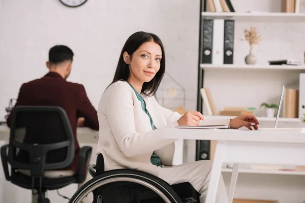 Smiling disabled businesswoman looking at camera while sitting in wheelchair at workplace — Stock Photo