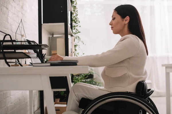 Attrayant femme d'affaires handicapée utilisant un ordinateur portable tout en étant assis en fauteuil roulant sur le lieu de travail — Photo de stock