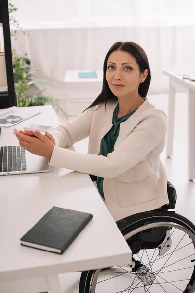 Bella donna d'affari disabili sorridente alla macchina fotografica mentre seduto sul posto di lavoro in sedia a rotelle — Foto stock