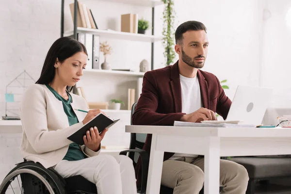 Attractive disabled businesswoman writing in notebook while sitting near colleague using laptop — Stock Photo