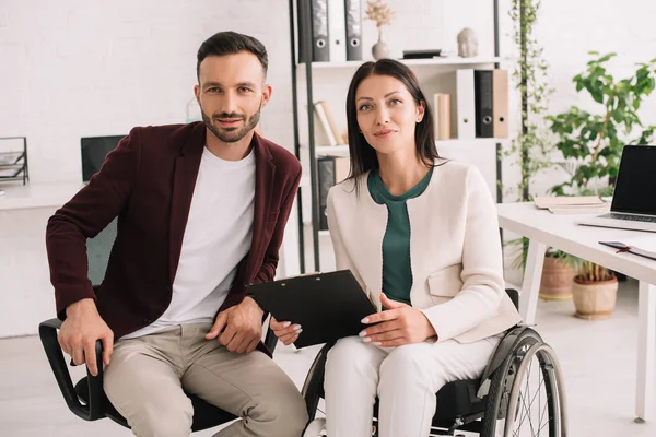 Mujer de negocios discapacitada positiva sosteniendo portapapeles mientras está sentada cerca de un socio de negocios en la oficina - foto de stock