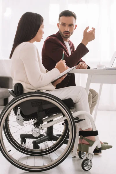 Mulher de negócios com deficiência segurando lápis e notebook enquanto conversa com o parceiro de negócios no escritório — Fotografia de Stock