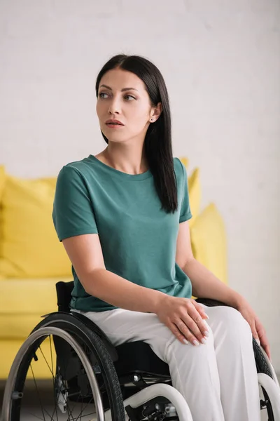 Серйозна жінка з обмеженими можливостями, дивлячись далеко, сидячи в інвалідному візку вдома — стокове фото