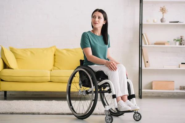 Mujer discapacitada positiva y sonriente mirando hacia otro lado mientras está sentada en silla de ruedas en casa - foto de stock