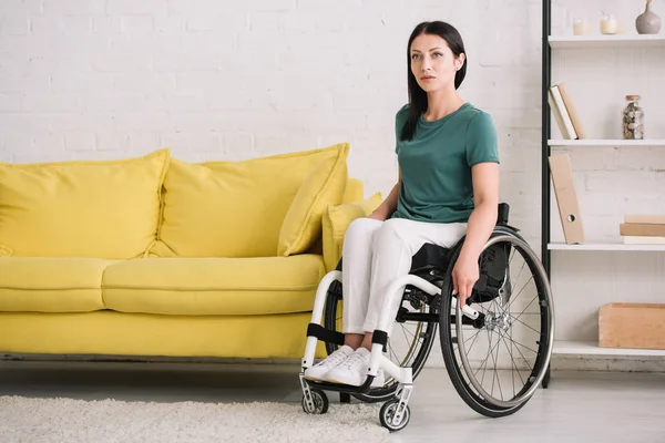 Mujer discapacitada joven y seria mirando hacia otro lado mientras está sentada en silla de ruedas en casa - foto de stock
