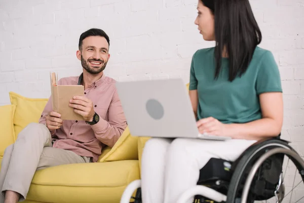 Pretty, disabled woman using laptop near cheerful boyfriend sitting on sofa with book — Stock Photo