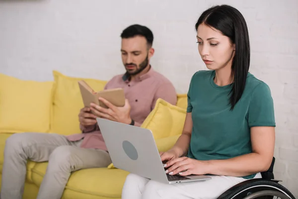 Concentrated disabled woman using laptop near boyfriend sitting on sofa and reading book — Stock Photo