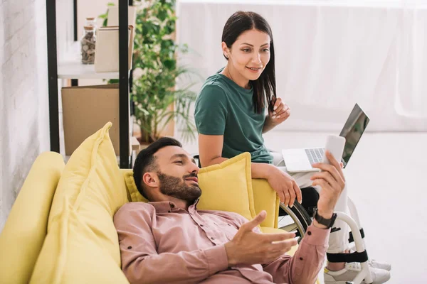 Fröhlicher Mann liegt mit Smartphone neben attraktiver behinderter Freundin auf Sofa und benutzt Laptop — Stockfoto