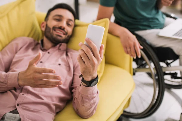 Homem alegre deitado no sofá com smartphone perto de namorada com deficiência usando laptop — Fotografia de Stock