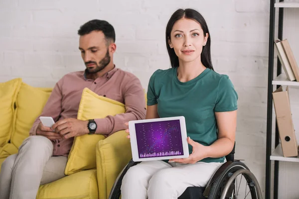 Mujer discapacitada sonriente mostrando tableta digital con infografías en la pantalla mientras está sentada cerca de novio usando teléfono inteligente - foto de stock