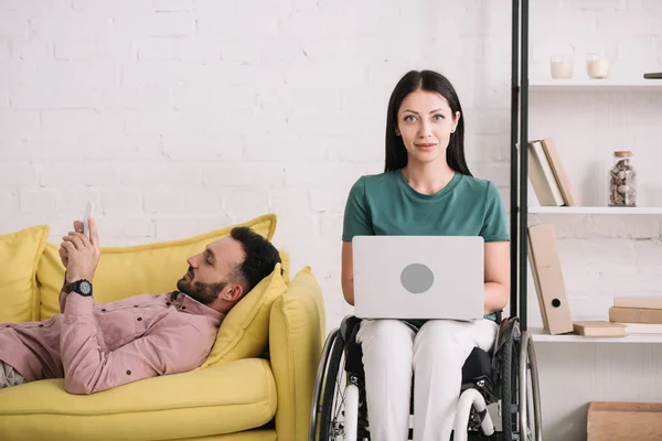 Smiling disabled woman using laptop while sitting near boyfriend lying on sofa and using smartphone — Stock Photo