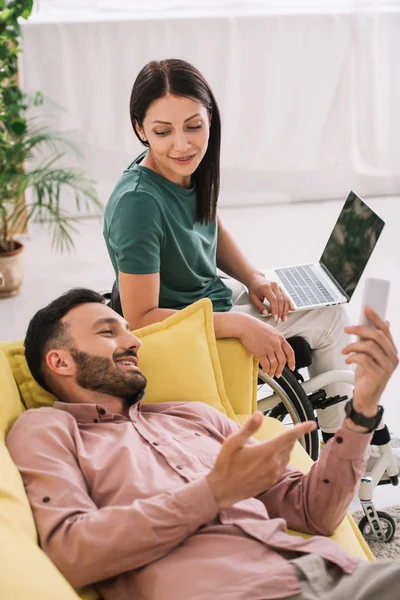 Sorrindo mulher deficiente usando laptop enquanto sentado perto namorado deitado no sofá com smartphone — Fotografia de Stock