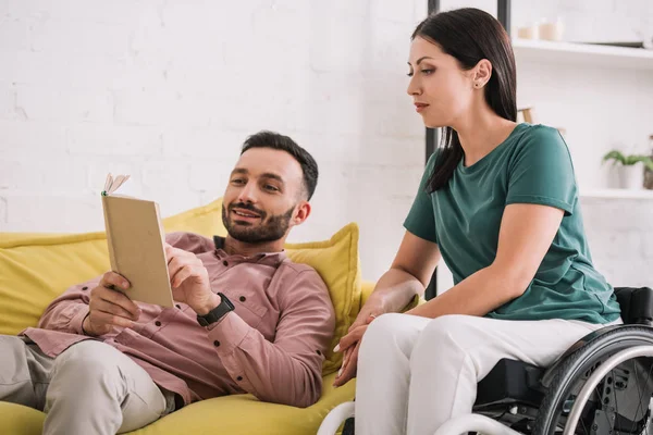 Junge behinderte Frau sitzt neben gutaussehendem Freund auf Sofa und liest Buch — Stockfoto